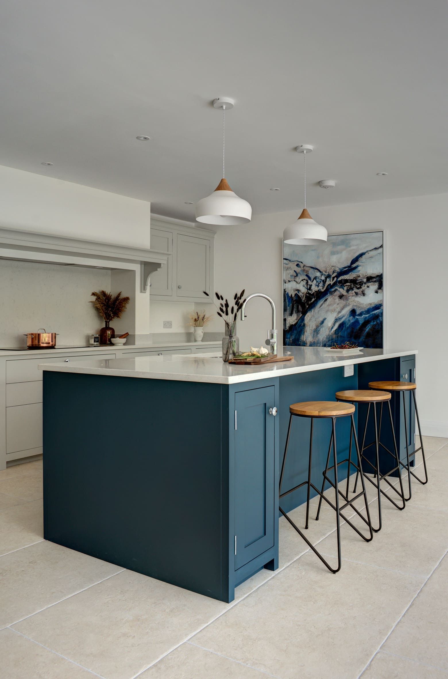 A modern bespoke kitchen design featuring a central island with a contrasting dark blue base and a white countertop. Three wooden bar stools with black metal legs are placed at the island. 