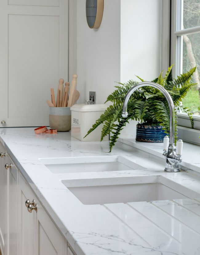 A built-in, bespoke kitchen sink is in the foreground, with a large green plant in a short blue vase sits in the window cill behind it.