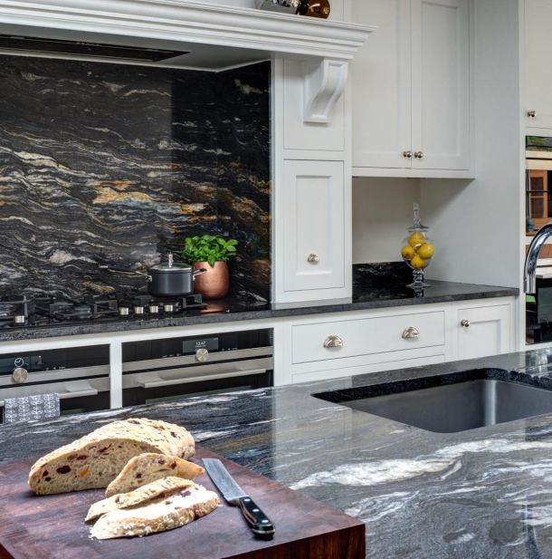 A loaf of bread, partially sliced on a bread board sits atop a bespoke, traditional granite kitchen top with a built-in sink basin. White cabinets contrast the dark colour of the worktop.