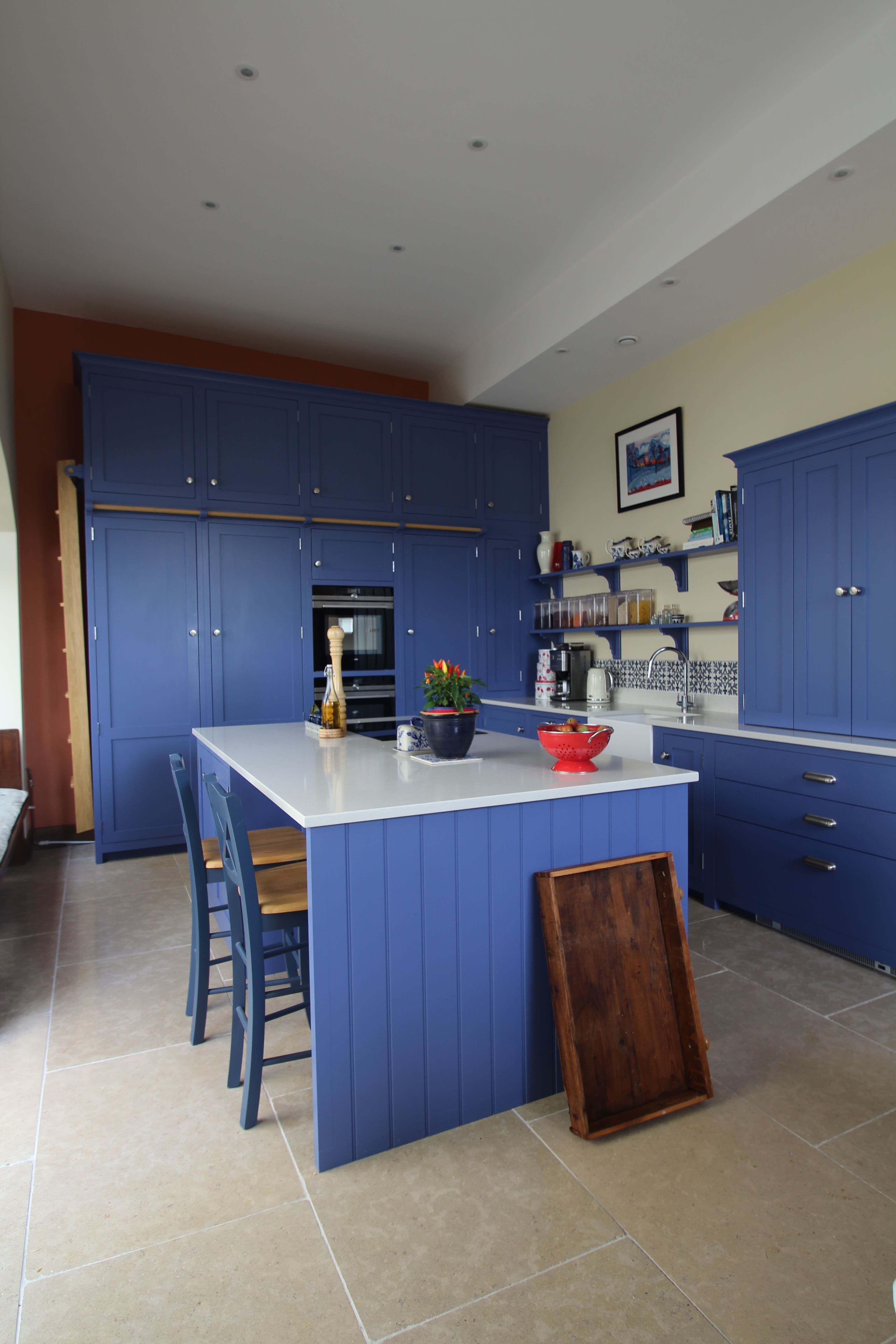 This is a vibrant kitchen with striking blue cabinetry and a matching central island, complemented by white worktops. The island also serves as a breakfast bar with two high blue stools. Natural light floods the space, reflecting off the light stone-tiled floor. 