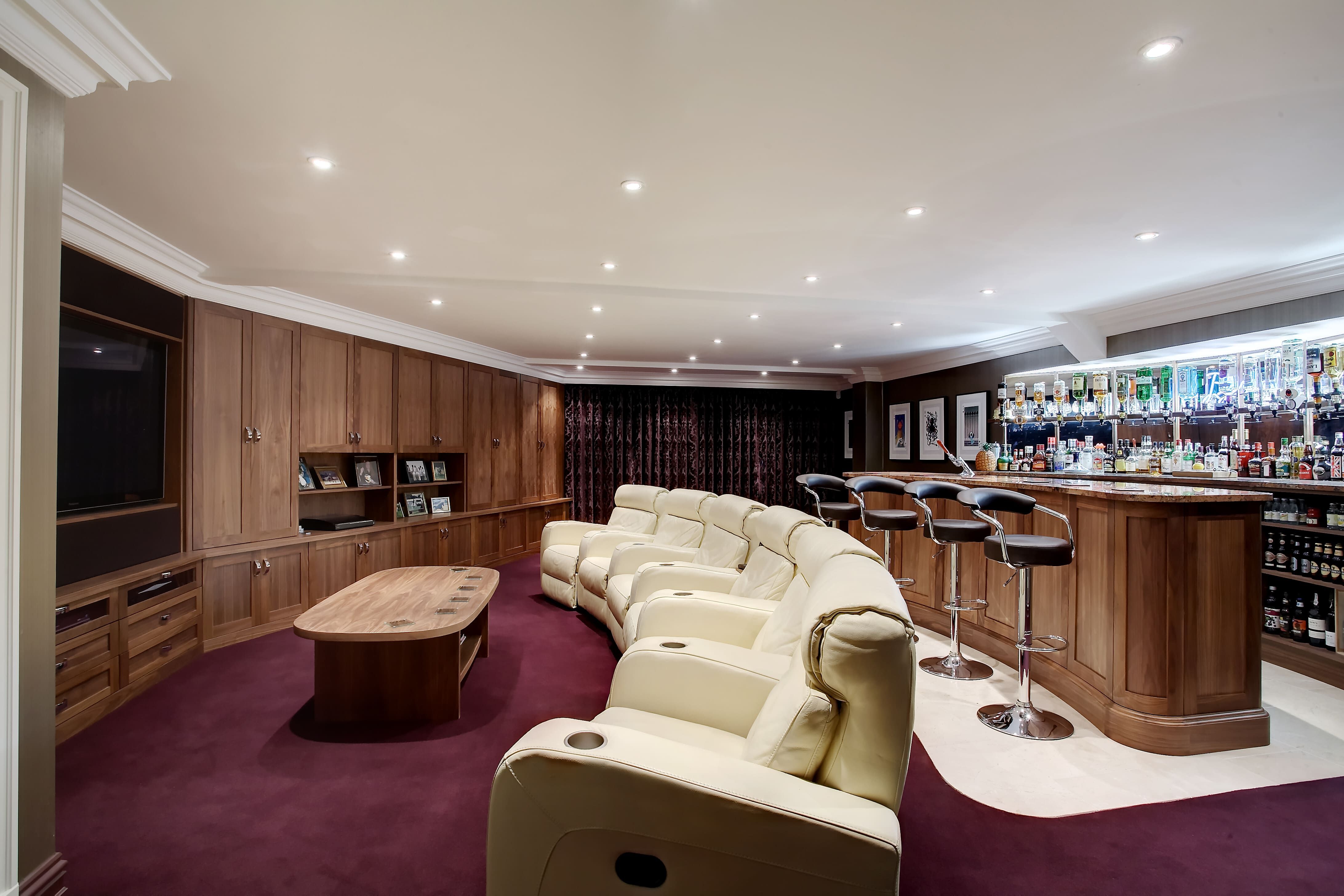 A bespoke, personal cinema room with bold colours. A burgundy carpet leads to a series of cream-coloured leather chairs in a radial shape. The room features floor-to-ceiling wooden cabinets, with a bar across the back wall.