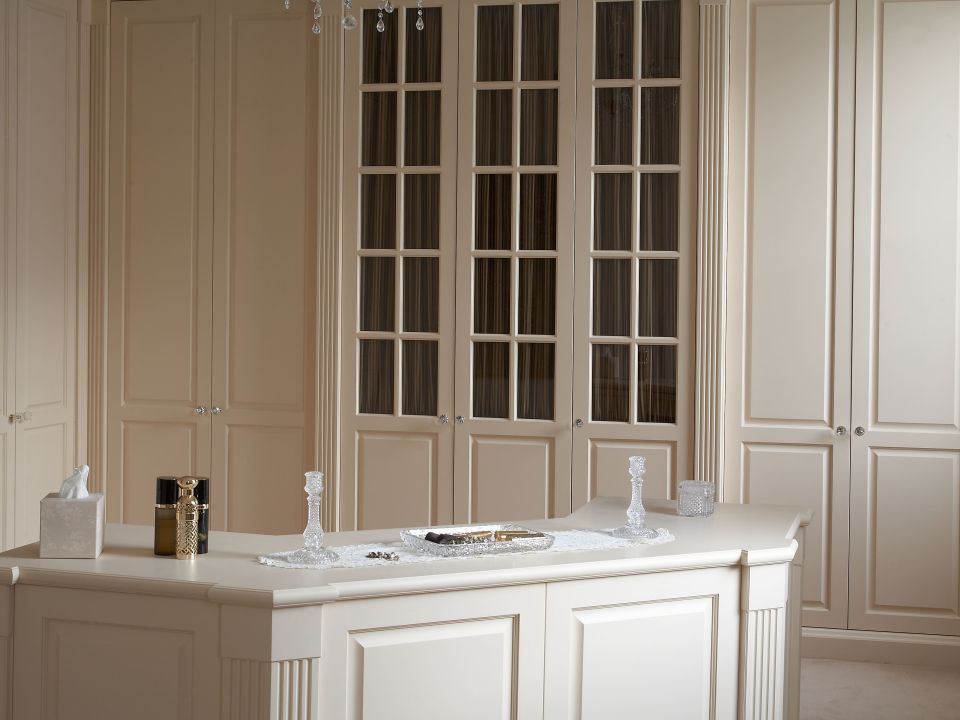 The room has cream fitted wardrobes with paneled doors and glass inserts, offering plenty of storage space. An elaborate crystal chandelier hangs from the ceiling, adding a touch of elegance.