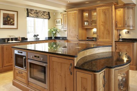 A traditional bespoke kitchen with natural wood cabinetry, black granite countertops, and built-in stainless steel appliances, complemented by under-cabinet lighting and a view to the garden.