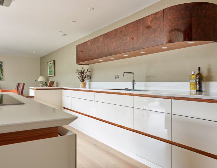 A modern bespoke kitchen with white high-gloss cabinetry, walnut accents, white countertops, and a unique burl wood extractor hood, all complemented by warm oak flooring and subtle recessed lighting.