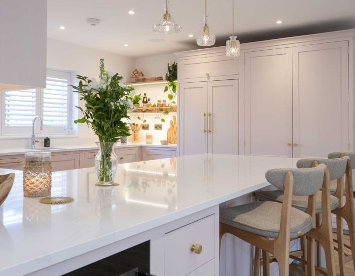 A light and elegant bespoke kitchen with blush pink cabinetry, gold hardware, white countertops, a central island with bar seating, and decorative pendant lights, enhanced by natural light and wooden accents.