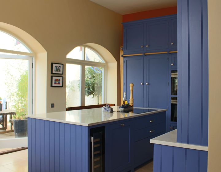 A striking blue, modular shaker kitchen with white counter tops is drowned in natural light from 2 large arched windows.