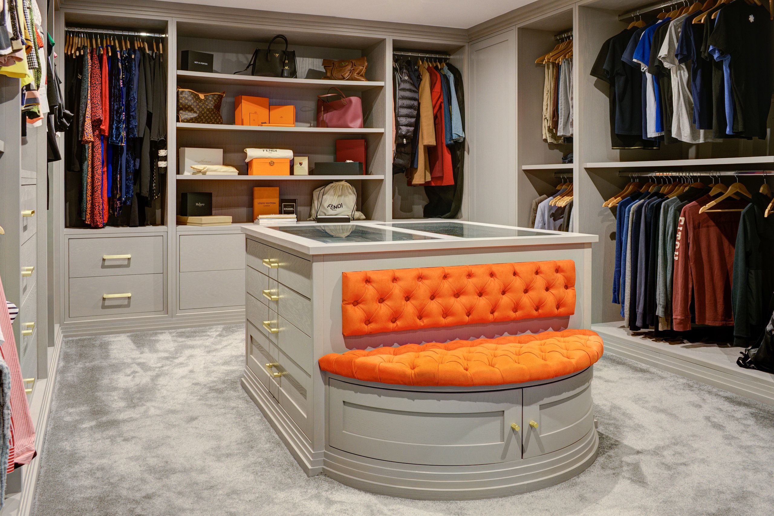 A bespoke dressing room with integrated cabinets surrounding the room. In the centre is a counter, with a striking orange-cushioned ottoman.