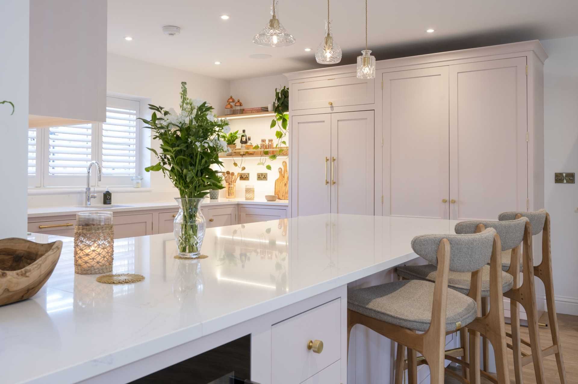 A light and elegant bespoke kitchen with blush pink cabinetry, gold hardware, white countertops, a central island with bar seating, and decorative pendant lights, enhanced by natural light and wooden accents.