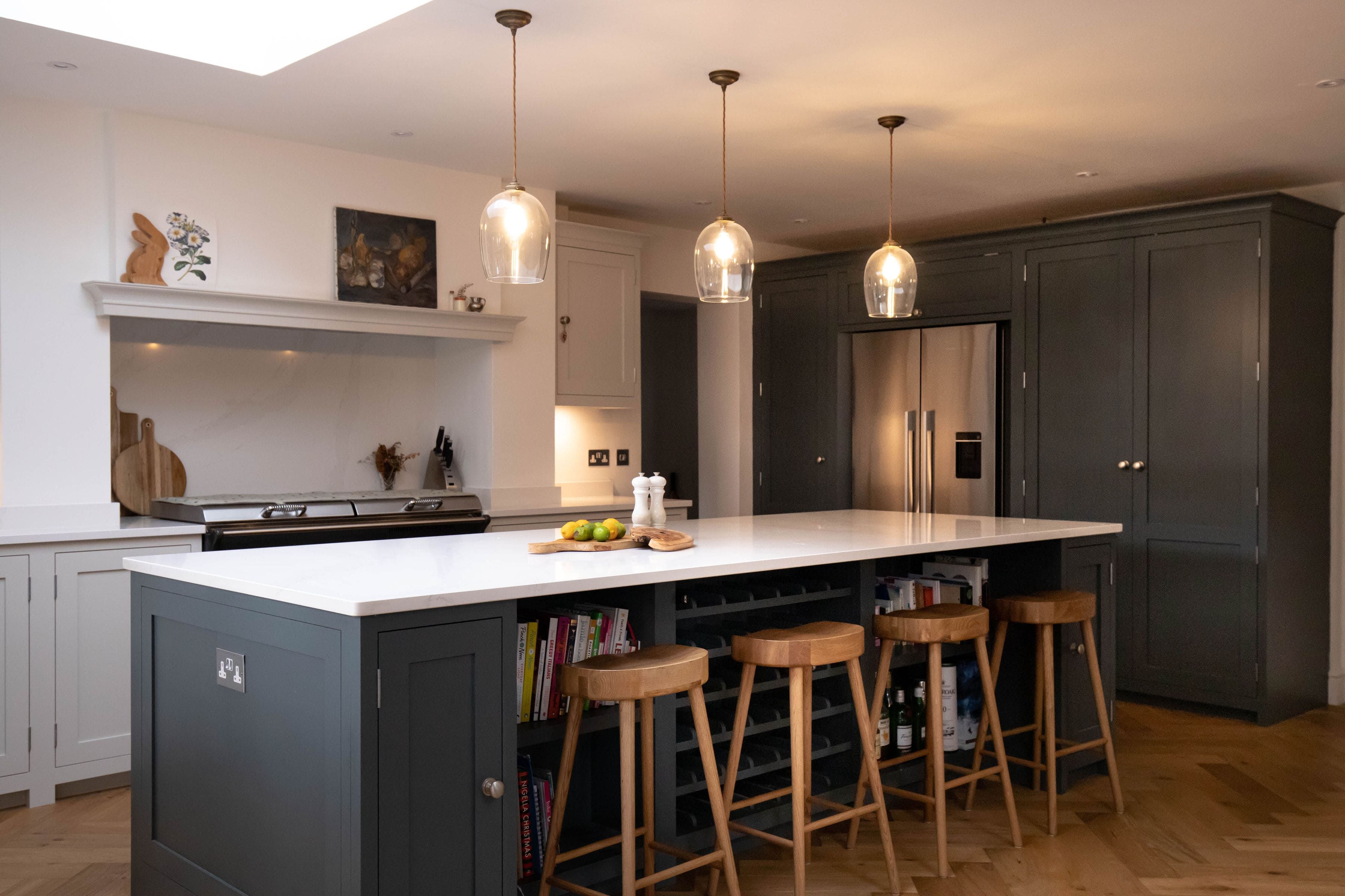 A large, bespoke kitchen island with dark blue cabinets and a white countertop sits in warm light from some hanging overhead lights. Varnished, herringbone-tiled wooden flooring provides strong contrast to the island, and complements the warming light of the room.