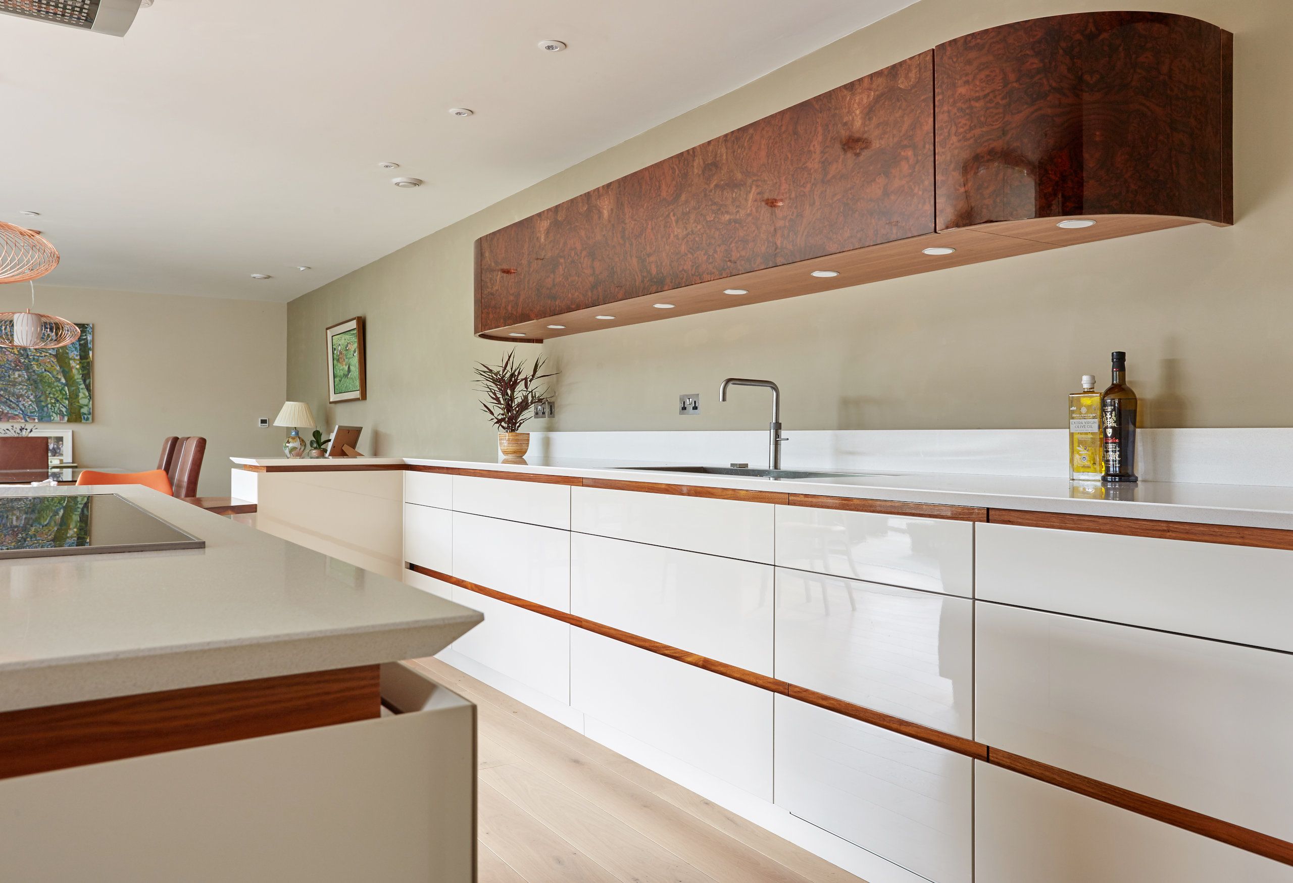 A modern bespoke kitchen with white high-gloss cabinetry, walnut accents, white countertops, and a unique burl wood extractor hood, all complemented by warm oak flooring and subtle recessed lighting.