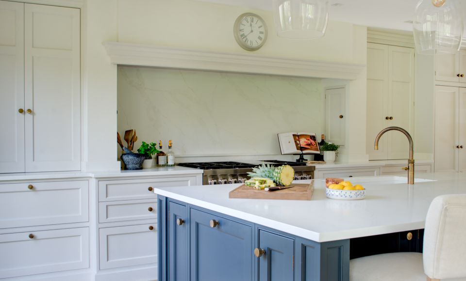 Modern bespoke traditional kitchen with white cabinets, marble splashback, and a central blue island under pendant lights.