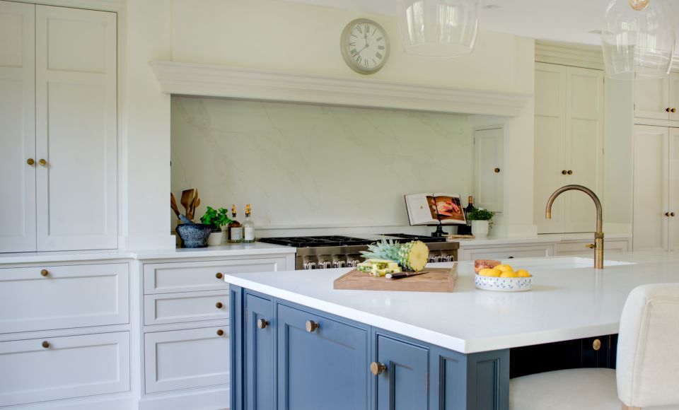 Modern bespoke traditional luxury kitchen with white cabinets, marble splashback, and a central blue island under pendant lights.