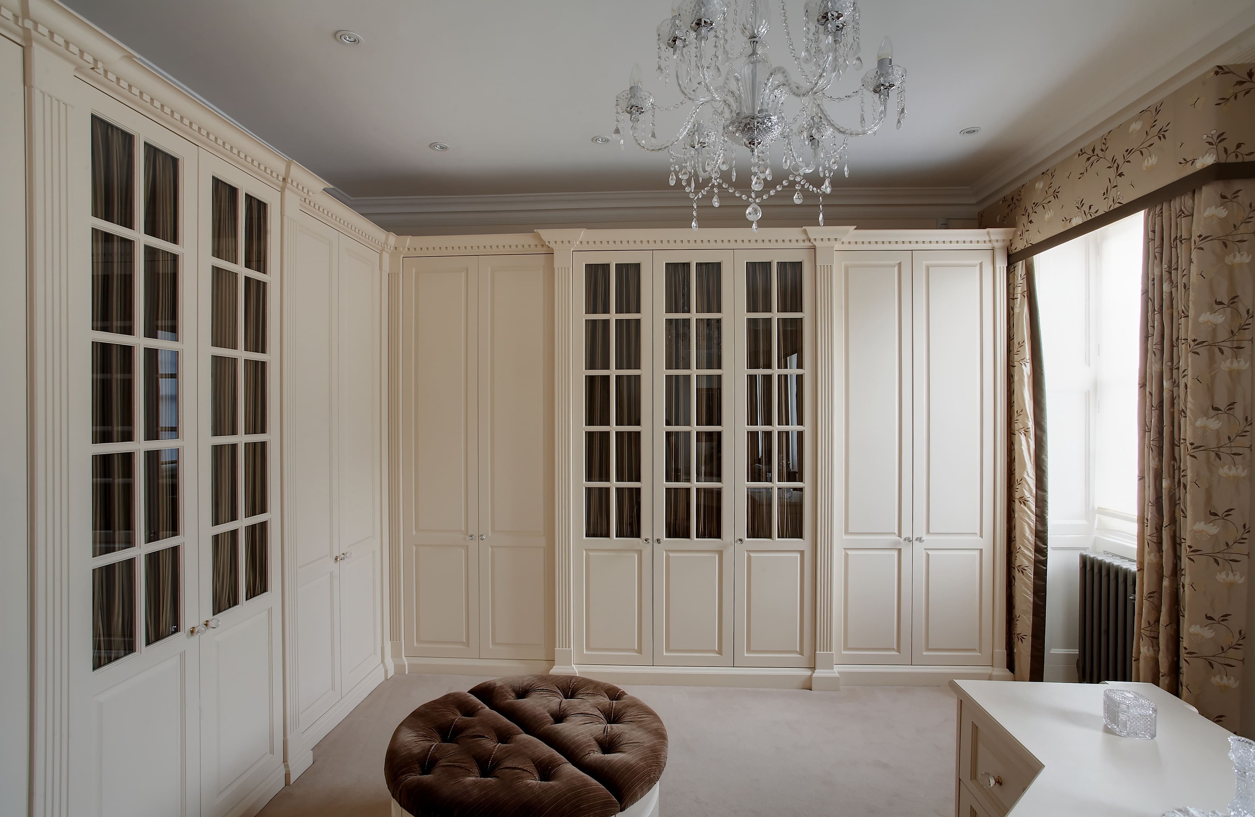 A luxurious bedroom with a neutral color palette, featuring a large, circular tufted ottoman in the center. The room has cream fitted wardrobes with paneled doors and glass inserts, offering plenty of storage space. An elaborate crystal chandelier hangs from the ceiling, adding a touch of elegance. 