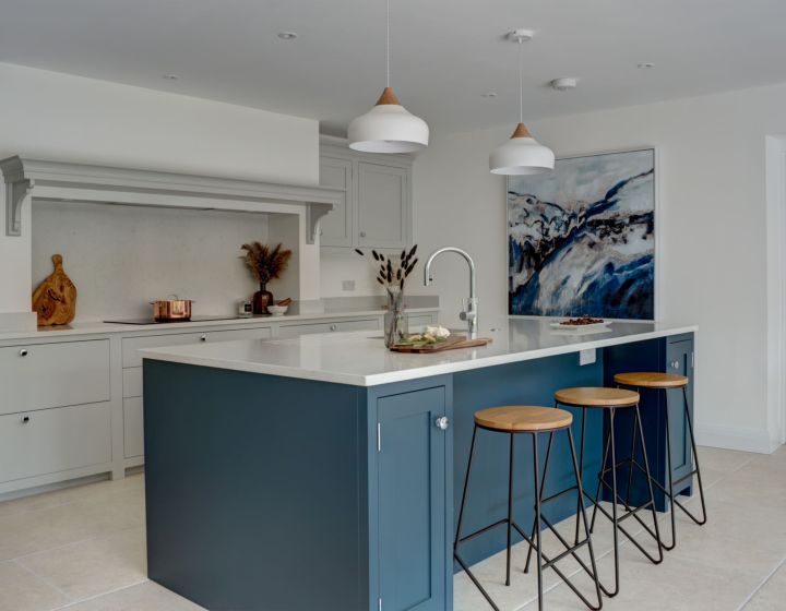 A contemporary bespoke kitchen with a two-tone design, featuring white cabinets, a teal island with a white countertop, wooden bar stools, pendant lighting, and decorative art on the wall, set on a light tiled floor.