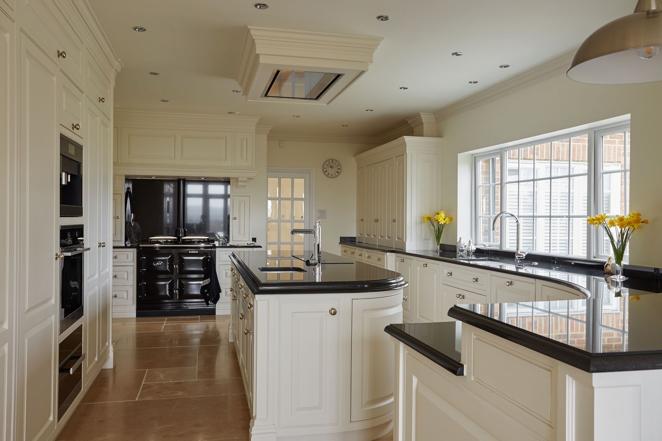 A refined bespoke kitchen with ivory cabinetry, black granite countertops, terracotta floor tiles, and a classic black range cooker, complemented by a skylight and stylish pendant lighting.