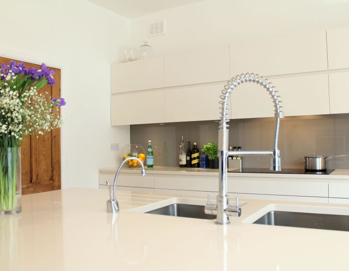 A bright, bespoke kitchen with high-gloss cream cabinets, a large central island with an integrated sink and professional chef's tap, accented by natural wood features and fresh flowers, under a backdrop of crisp white walls.