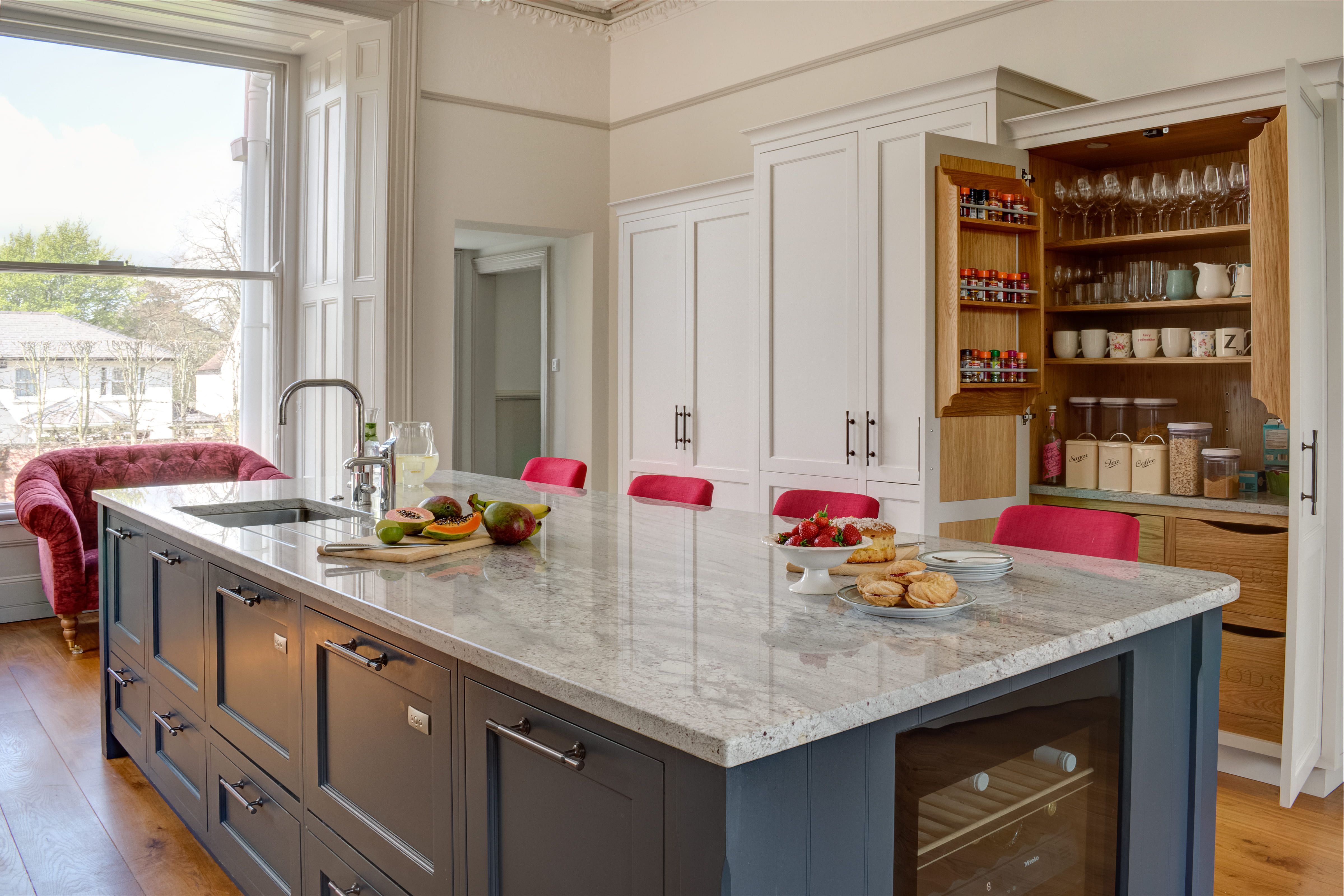 A large, navy blue, bespoke Shaker kitchen island with a grey marble worktop. A large red arm chair and red stools provide some strong contrast and bring more colour to the room. A built-in pantry is shown open in the background. 