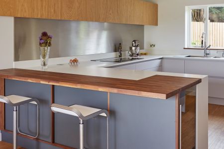 A sleek bespoke kitchen with charcoal grey cabinets, walnut worktops, a modern induction hob, stainless steel backsplash, and light oak flooring, accented by contemporary bar stools and pendant lighting.