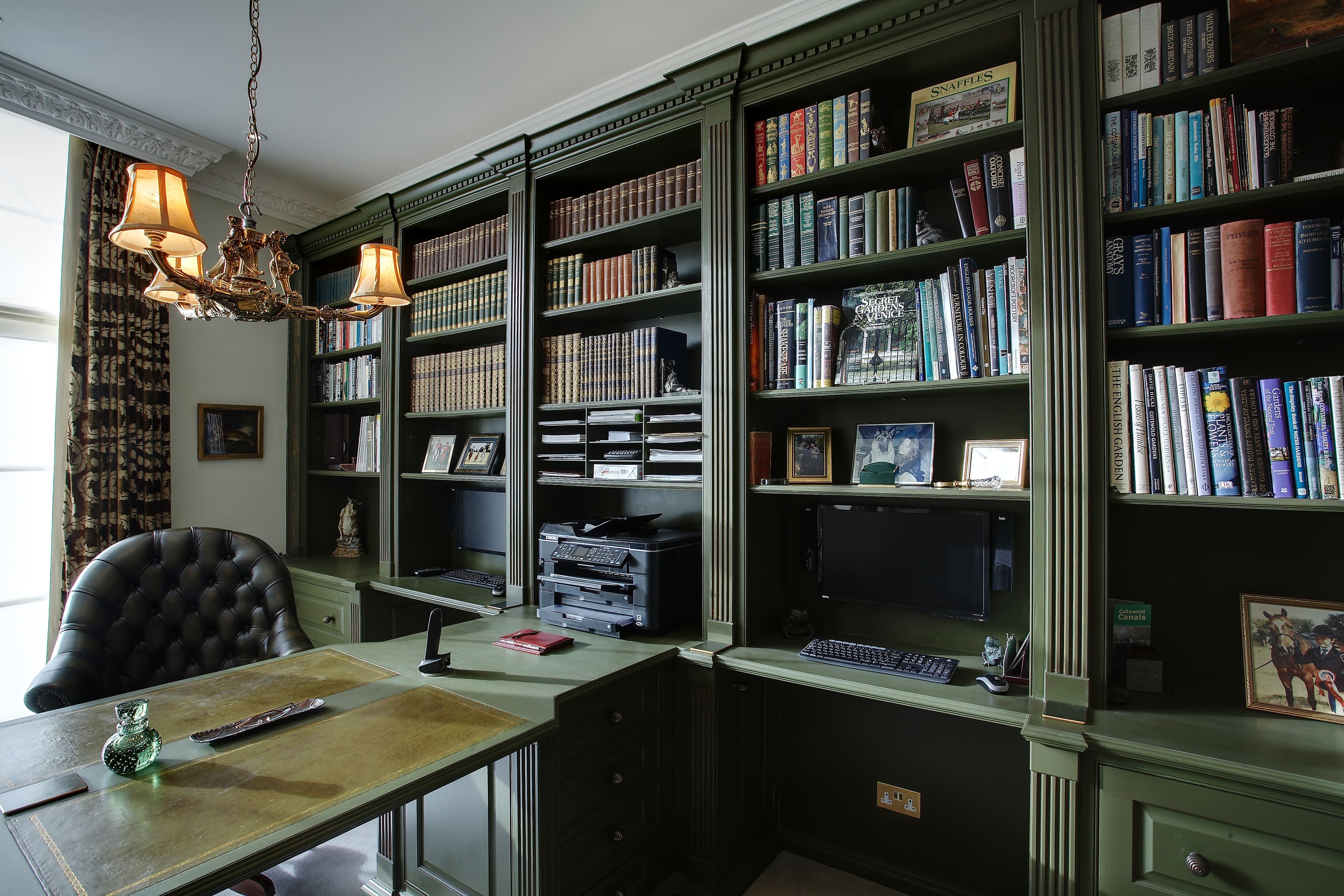 A sophisticated home study with dark green built-in bookshelves filled with a colourful array of books. In front of the bookshelves is a classic chest of drawers with a traditional design and dark metal handles.