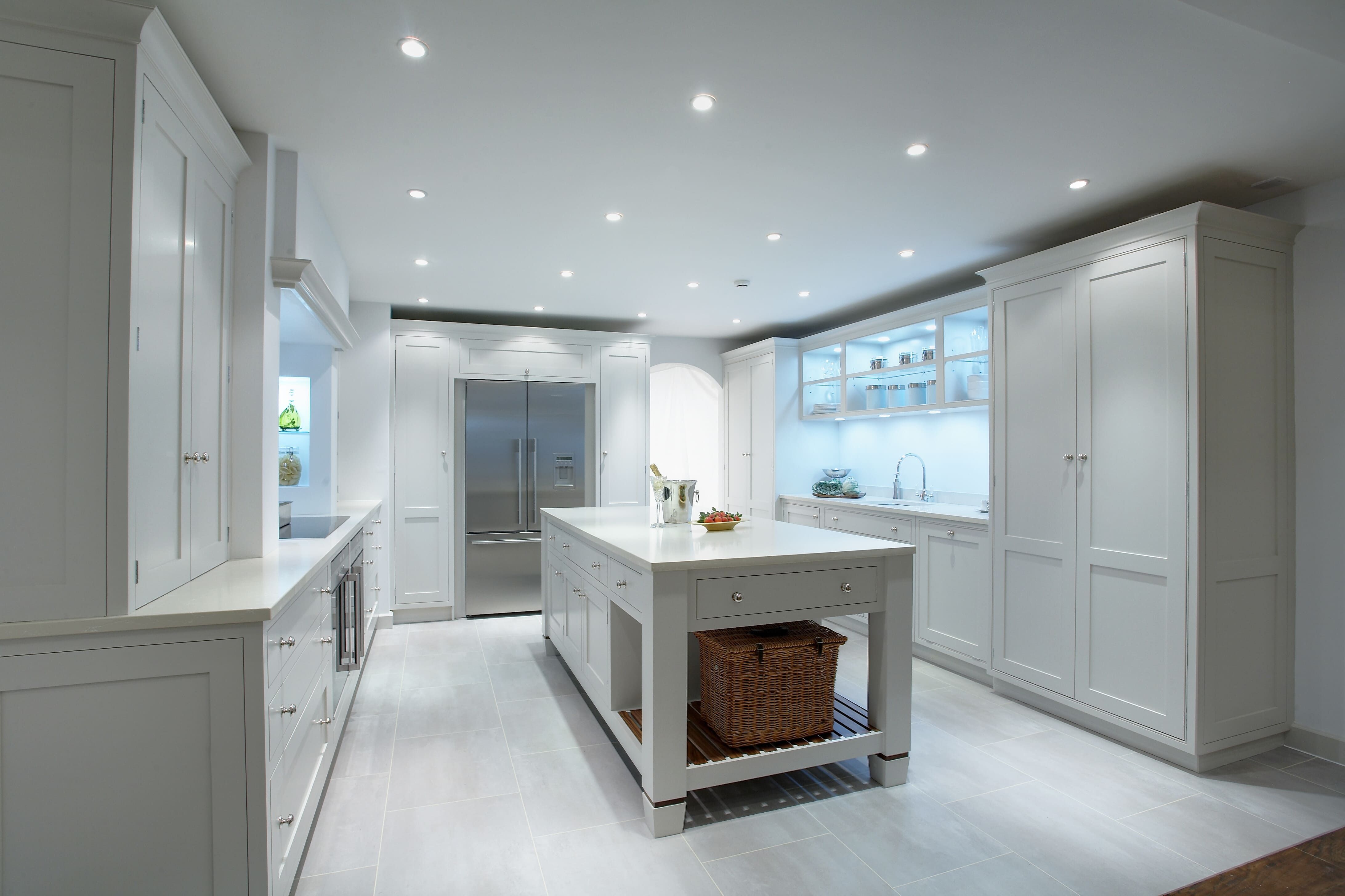 A large, light-grey bespoke kitchen and island is bathed in a cool light from a series of built-in overhead spot lights. A large fridge-freezer can be seen set in to a cabinet on the back wall.