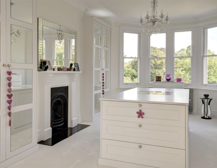 A white, bespoke dressing room, with large drawer set/worktop in the centre. Pink hanging decorations provide a contrasting colour.