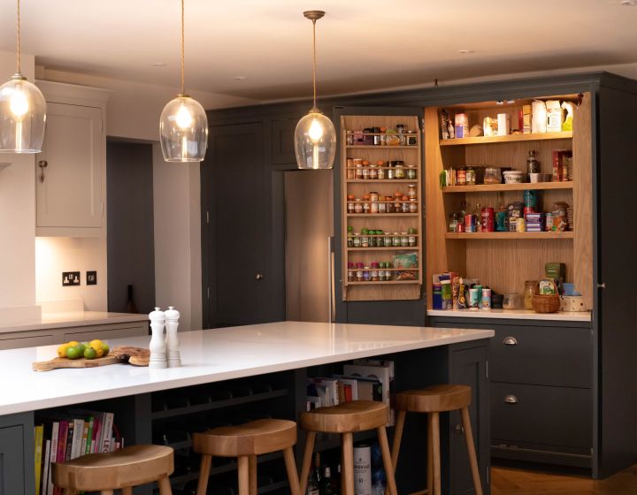 A warm, bespoke kitchen with dark grey cabinetry, a white quartz island, wooden bar stools, and glass pendant lights, featuring an open pantry with neatly organized shelves.