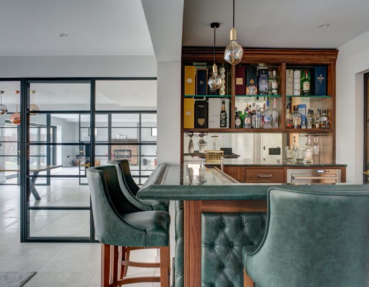 A bespoke personal bar, with green leather seating. A large game room can be seen through some floor-to-ceiling glass doors.