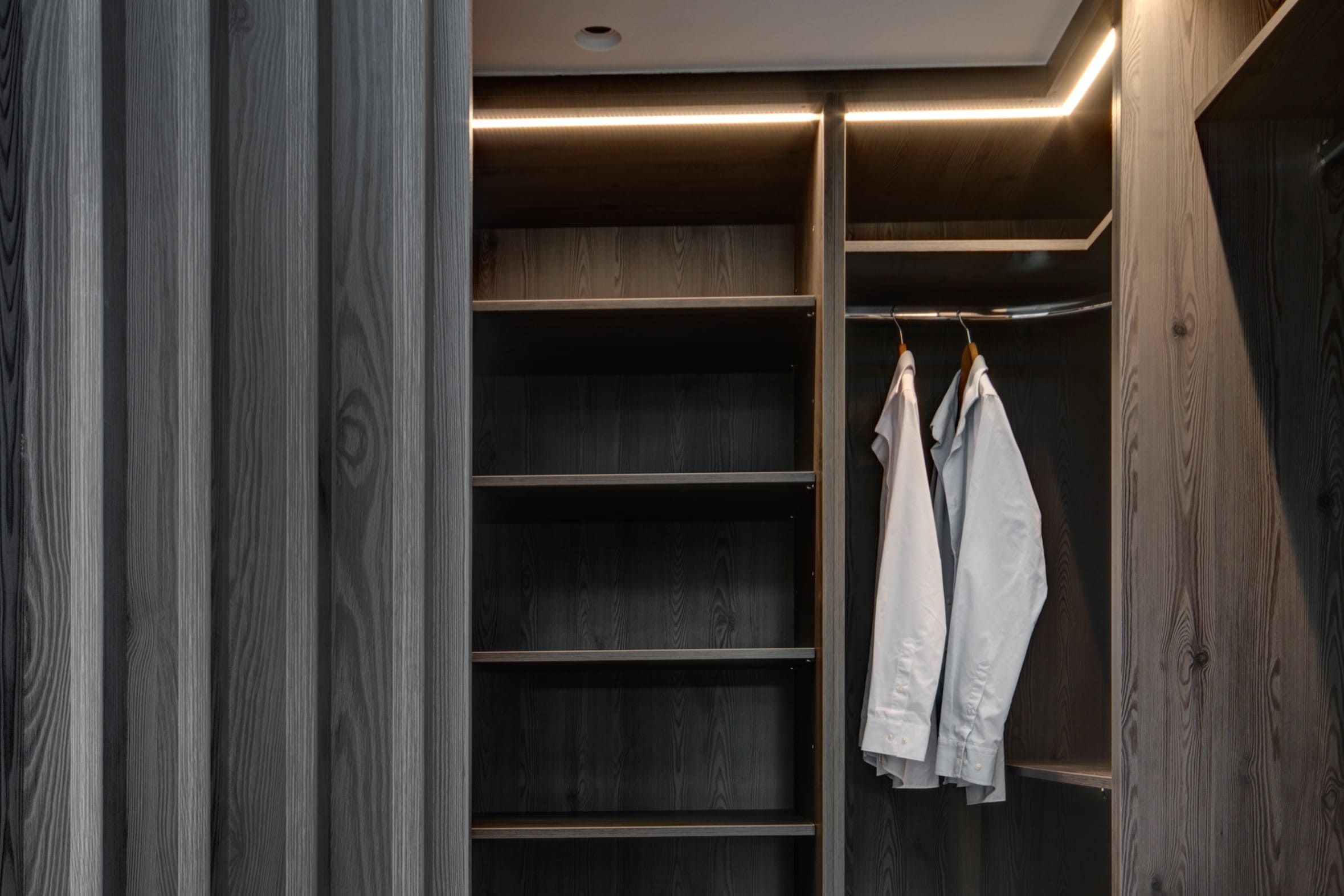 An integrated, walk-in bespoke dressing room. Soft lighting provides a warming tone to the grey wood cabinets.