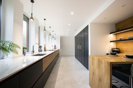 A modern bespoke kitchen with long, dark grey cabinets, white countertops, natural wood open shelving, and a wood-topped breakfast bar, illuminated by unique pendant lights and complemented by pale tiled flooring.
