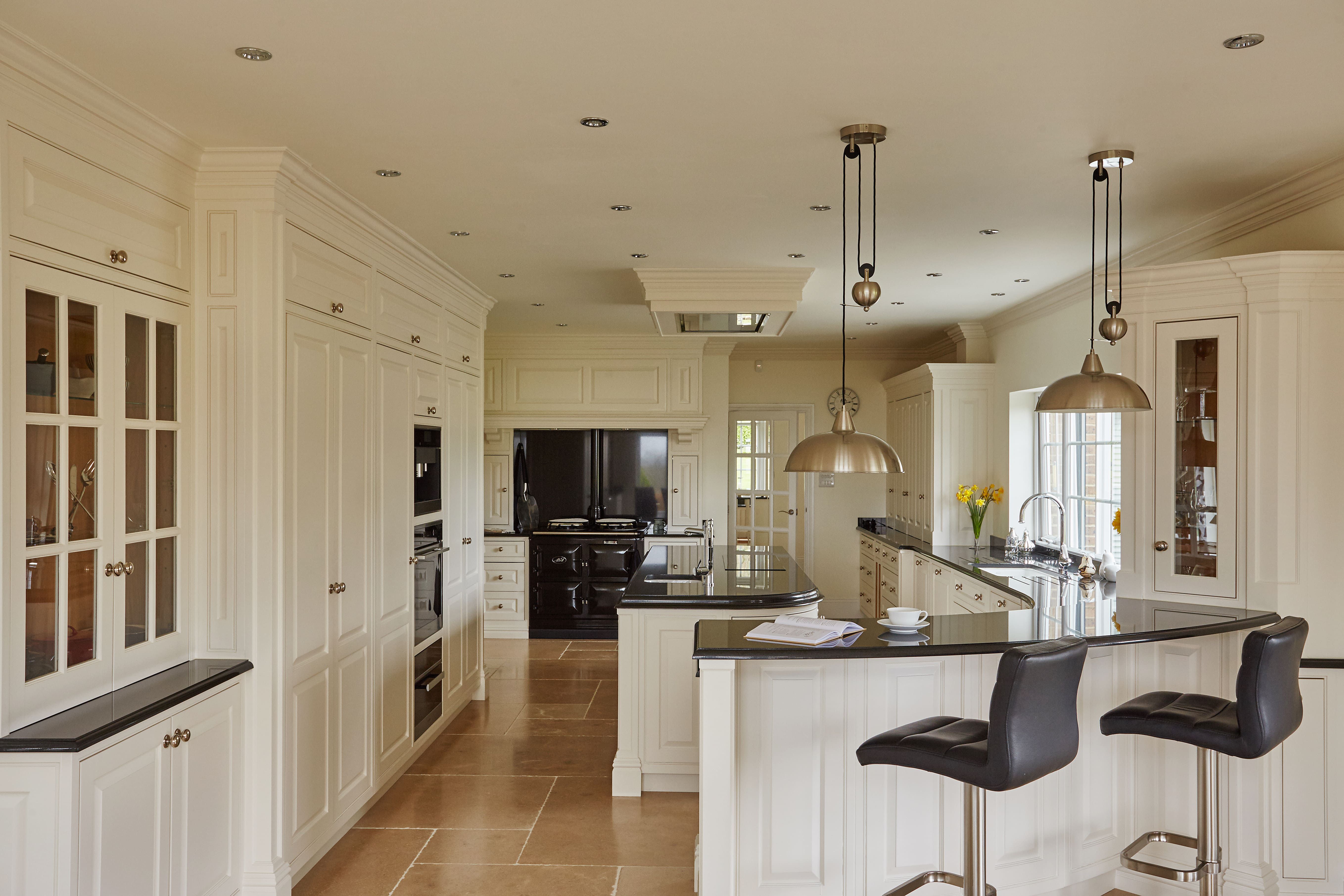 An expansive, classic kitchen with cream cabinetry and black countertops. The kitchen features glass-fronted cabinets, a large black stove, and built-in modern appliances. There's a central island with a built-in sink and a breakfast bar with two black leather bar stools.