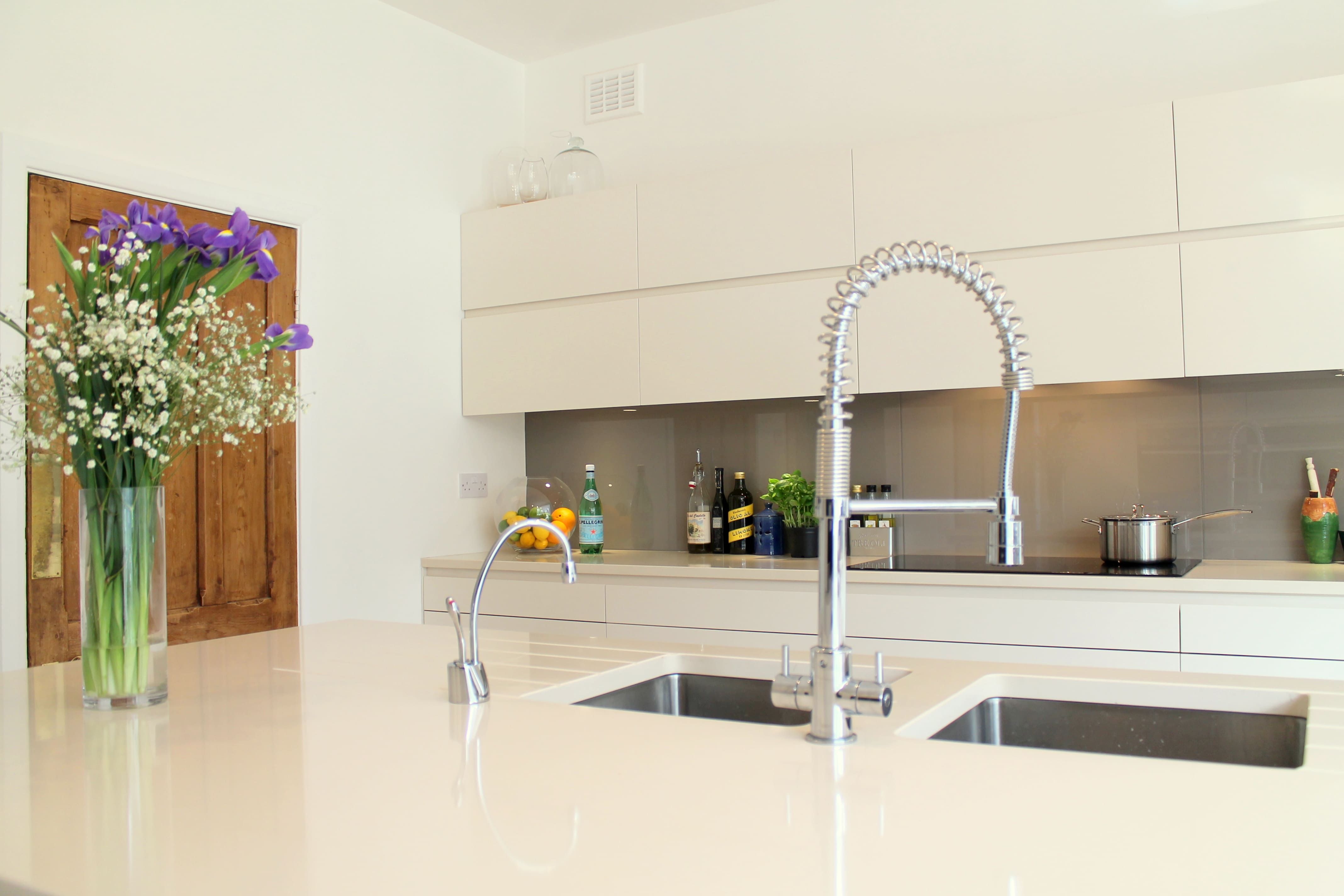 A bright, bespoke kitchen with high-gloss cream cabinets, a large central island with an integrated sink and professional chef's tap, accented by natural wood features and fresh flowers, under a backdrop of crisp white walls.