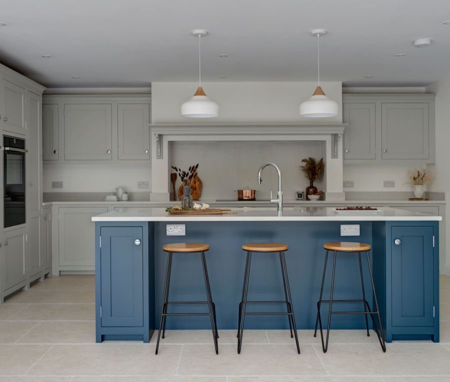 A strong blue, bespoke kitchen island contrasts the white walls and light grey cabinets of the rest of the bespoke kitchen. Some minimalist black stools with wooden seats are seen in the foreground next to the island.
