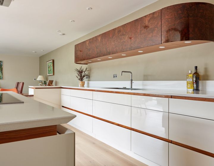 A modern bespoke kitchen with white high-gloss cabinetry, walnut accents, white countertops, and a unique burl wood extractor hood, all complemented by warm oak flooring and subtle recessed lighting.