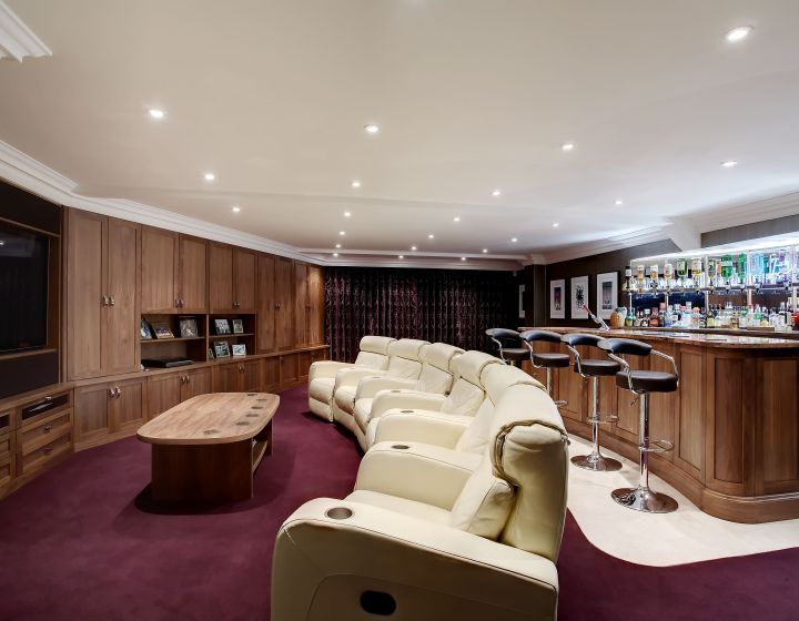 A bespoke, personal cinema room with bold colours. A burgundy carpet leads to a series of cream-coloured leather chairs in a radial shape. The room features floor-to-ceiling wooden cabinets, with a bar across the back wall.