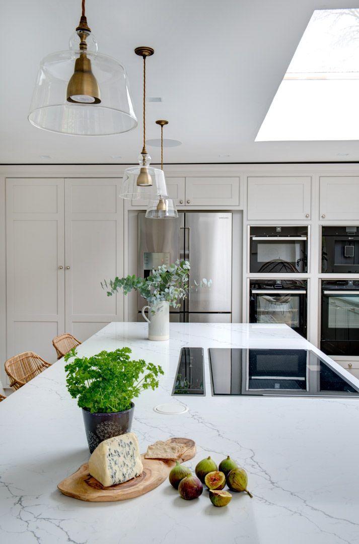  A modern and bright bespoke kitchen interior with white marble countertops and sleek cabinetry. Two pendant lights with a vintage design hang above the island. There is a large skylight providing natural light, enhancing the room's airy feel. 