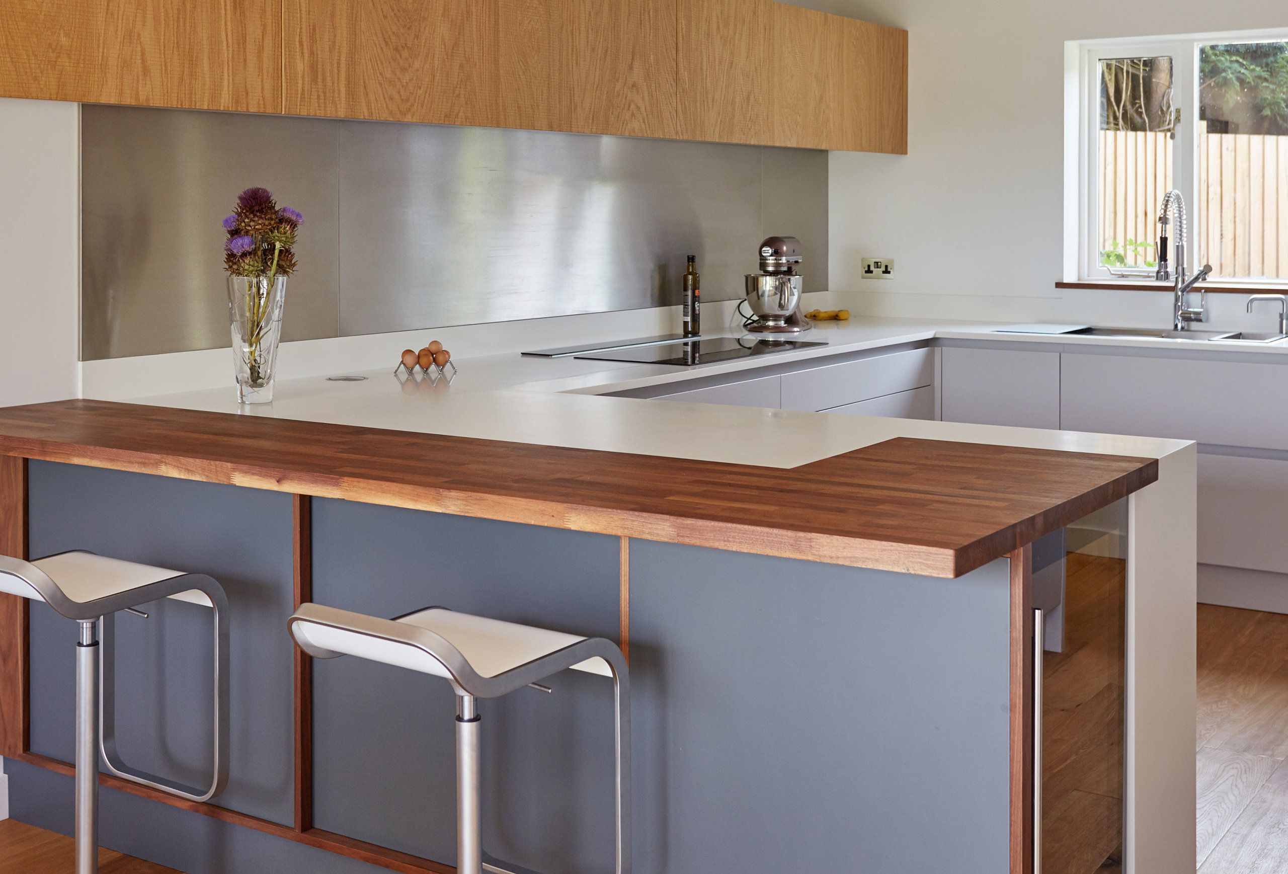 A sleek bespoke kitchen with charcoal grey cabinets, walnut worktops, a modern induction hob, stainless steel backsplash, and light oak flooring, accented by contemporary bar stools and pendant lighting.