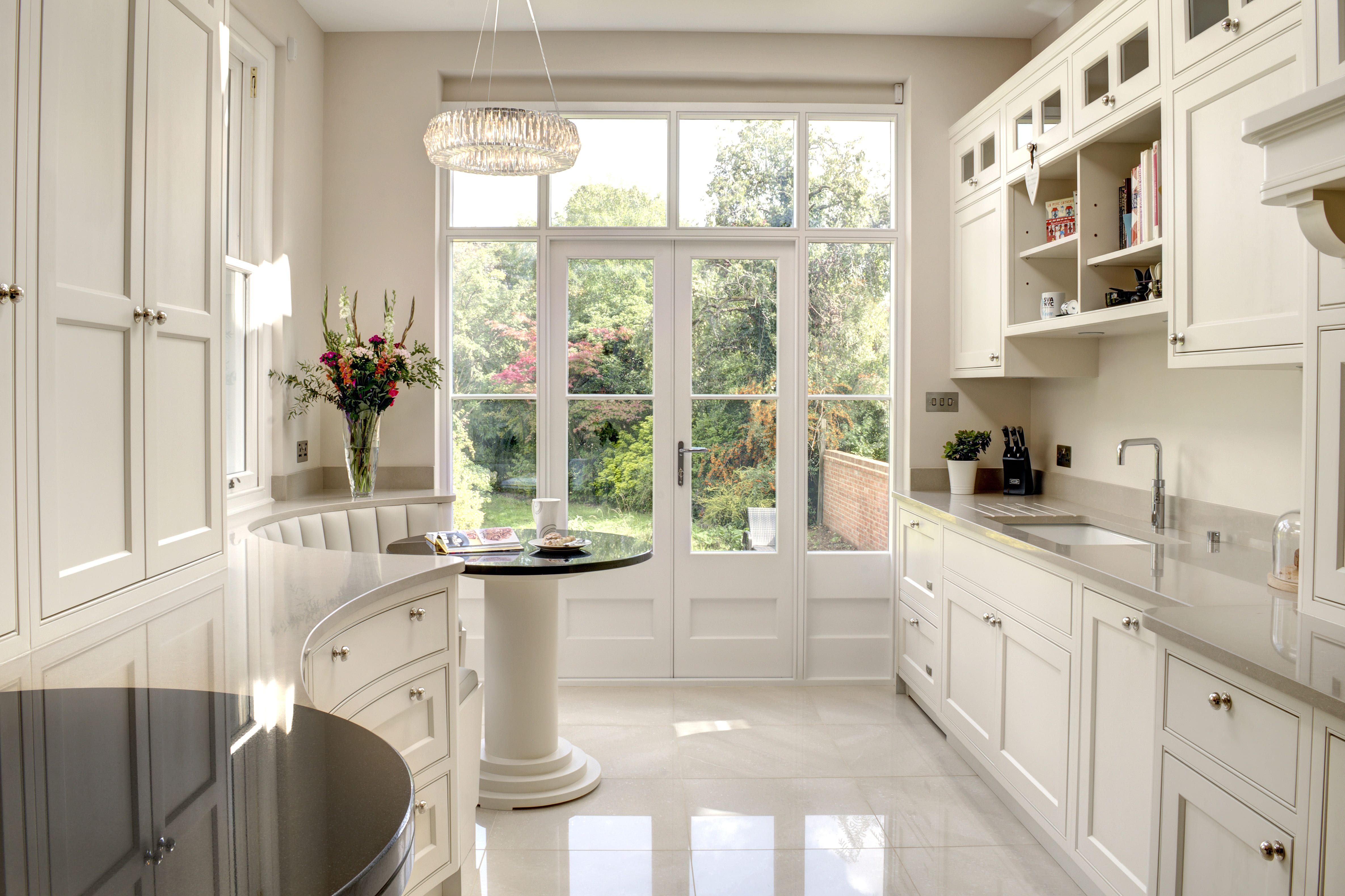 A cream-coloured bespoke Shaker kitchen, with a circular seating area with a round black table looks out some external doors to a colourful garden.