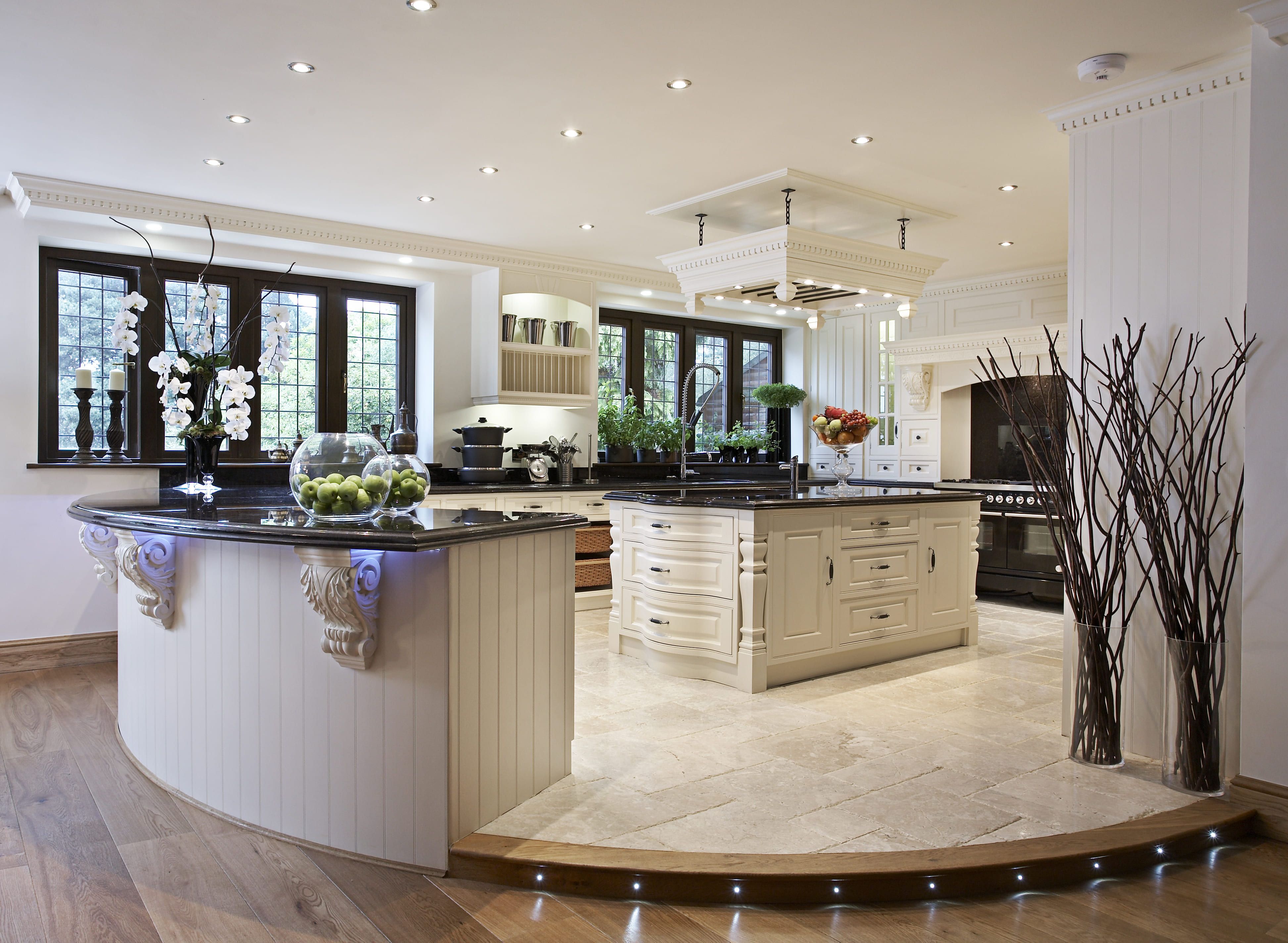 A spacious bespoke kitchen with cream panelled cabinetry, black granite worktops, a central island, and a traditional range cooker, all set against a backdrop of natural stone flooring and an ornate plastered ceiling.