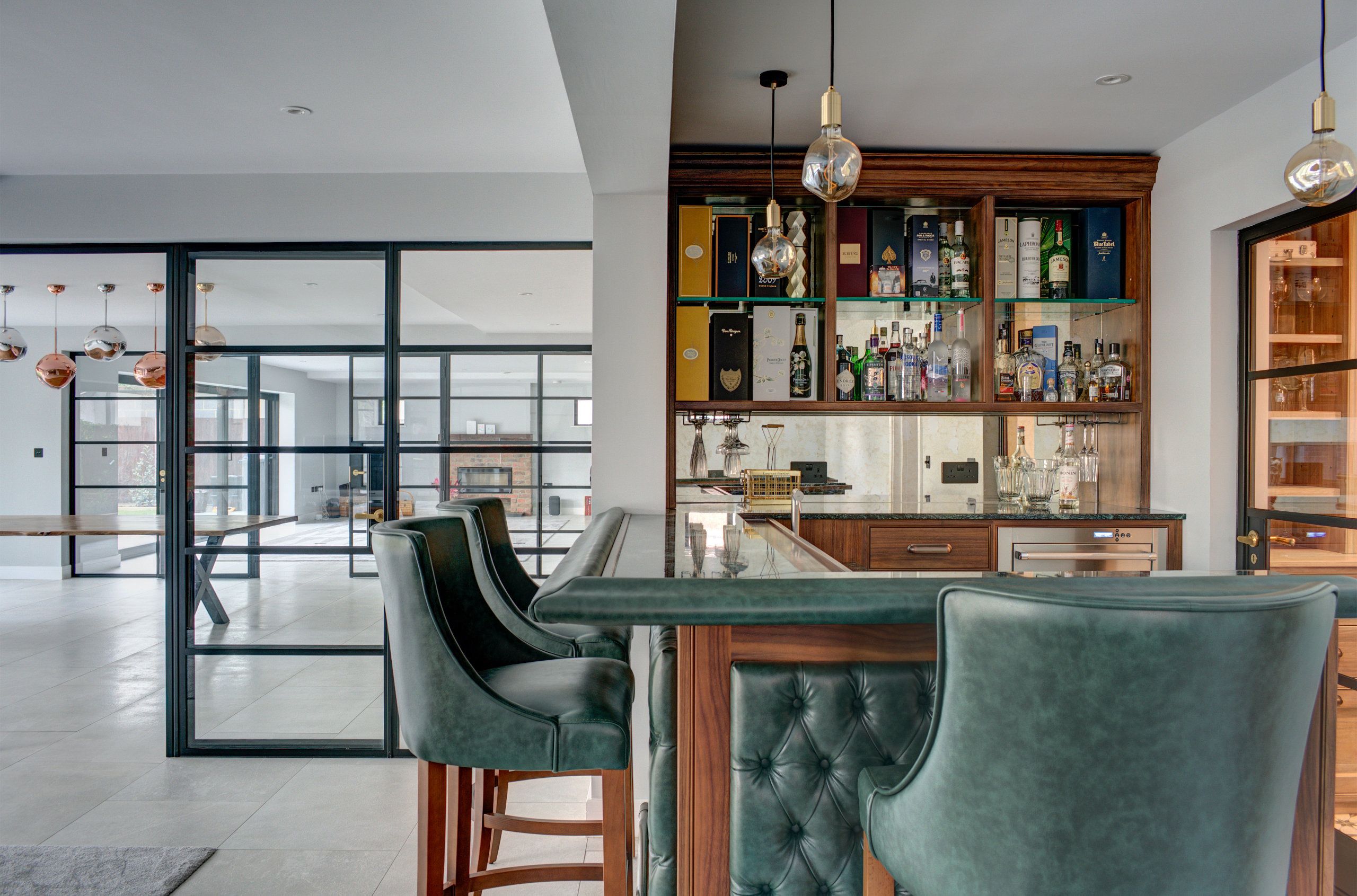 A bespoke personal bar, with green leather seating. A large game room can be seen through some floor-to-ceiling glass doors.