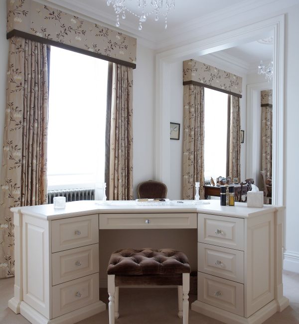 An elegant vanity area in a bedroom with cream-colored walls and a plush carpet. The vanity table is cream with a marble top and equipped with drawers. Above it hangs a luxurious crystal chandelier. A classic tufted stool sits in front of the vanity.