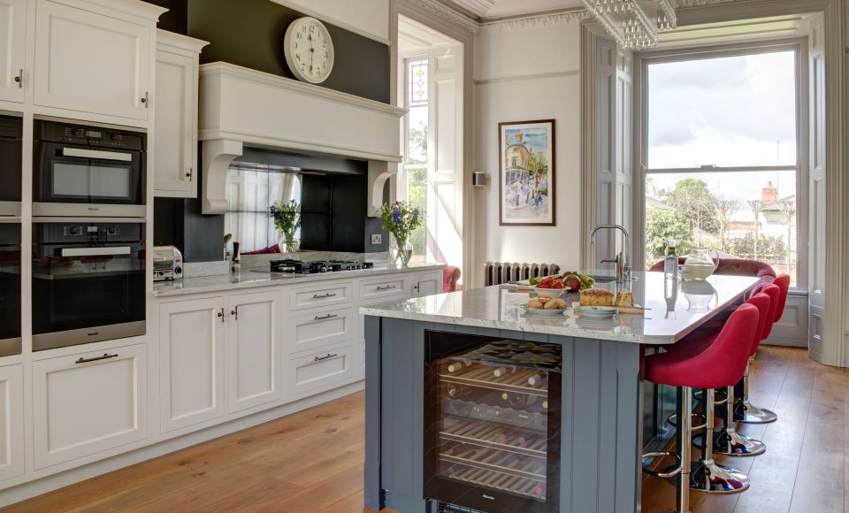 A bespoke shaker luxury kitchen with white cabinets, a grey island with red stools. A large, floor-to-ceiling window bathes the room in natural light.