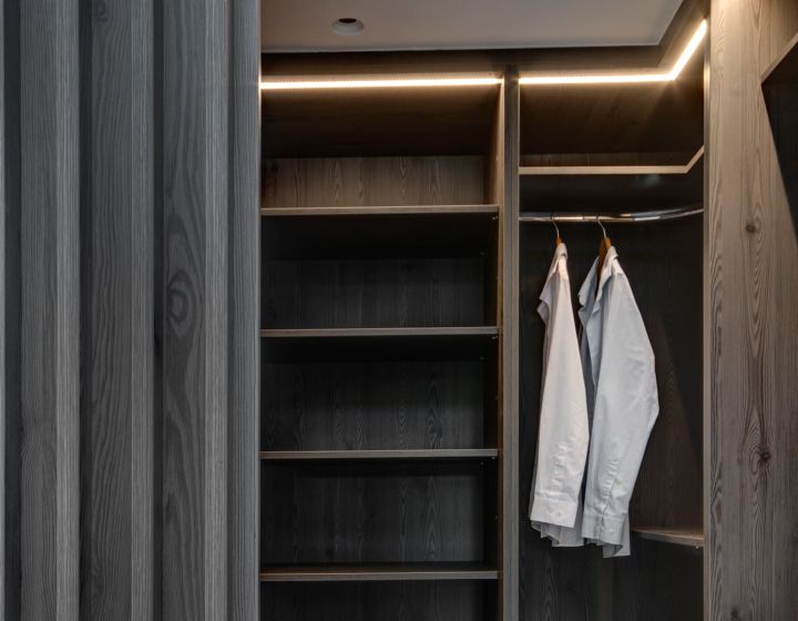An integrated, walk-in bespoke dressing room. Soft lighting provides a warming tone to the grey wood cabinets.
