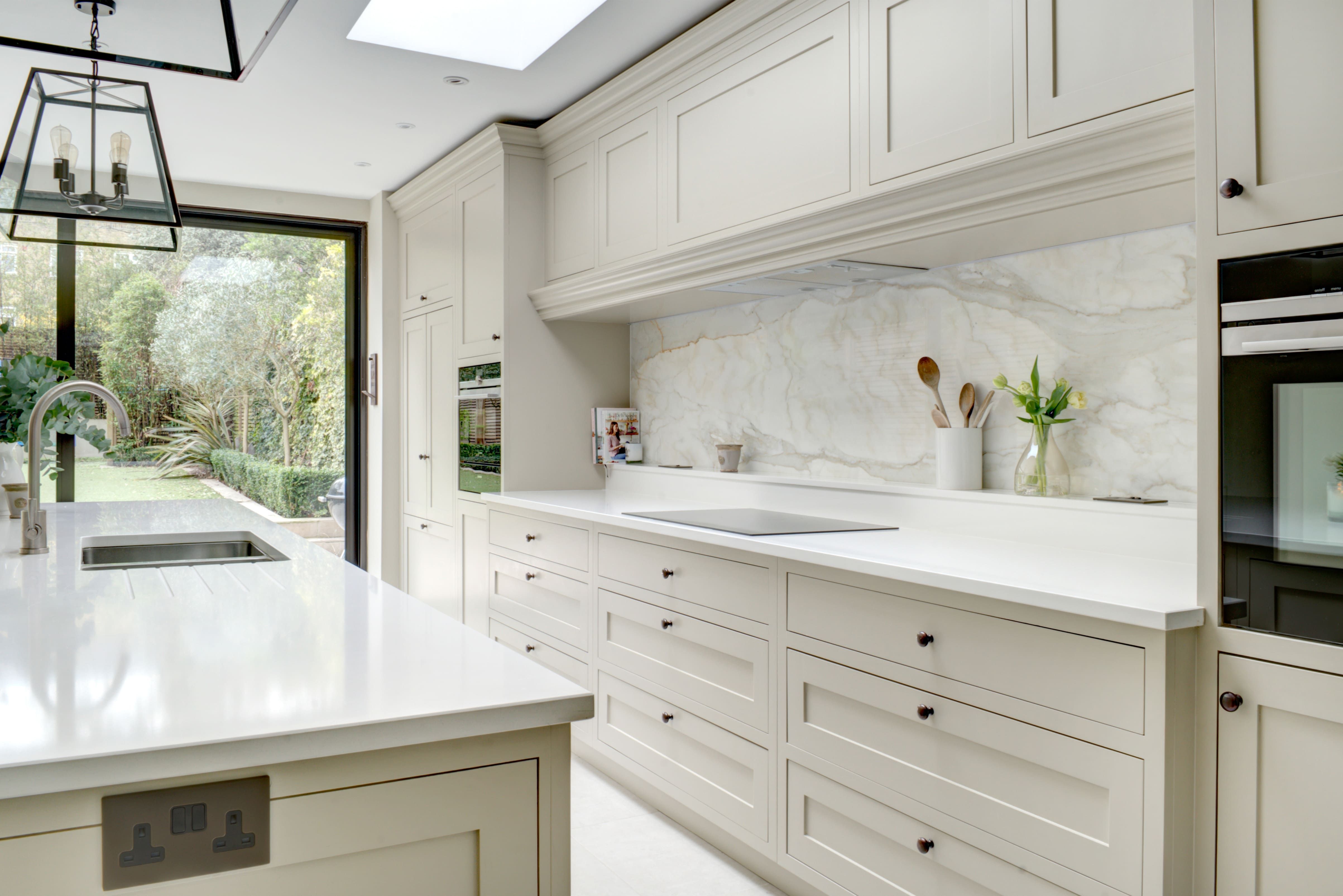A cream and white bespoke kitchen is bathed in natural light from a floor-to-ceiling patio door, opening to a large, well-kept garden.