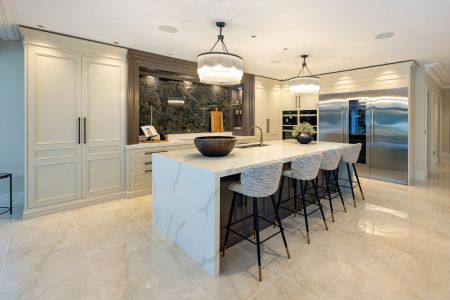 A bespoke traditional kitchen featuring a marble-topped island with bar stools, elegant twin pendant lights, and integrated high-end appliances, set against a backdrop of polished marble flooring.