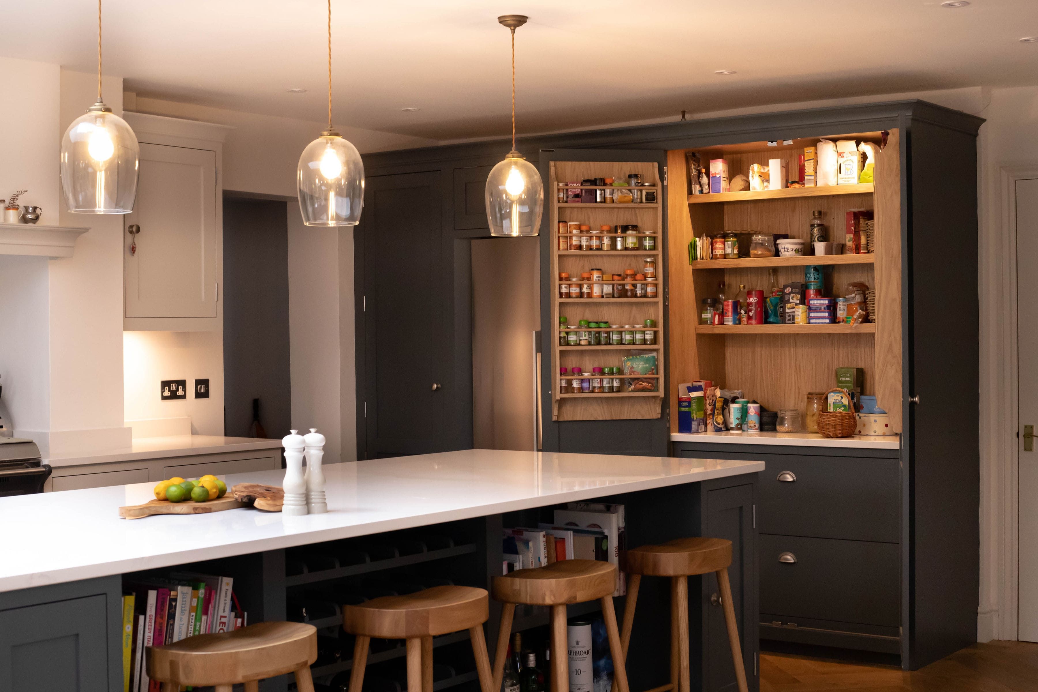 A warm, bespoke kitchen with dark grey cabinetry, a white quartz island, wooden bar stools, and glass pendant lights, featuring an open pantry with neatly organized shelves.