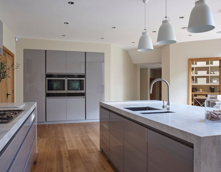 A minimalist bespoke kitchen with sleek grey cabinetry, a concrete effect island, natural wood flooring, and modern pendant lighting, creating a clean and airy space.