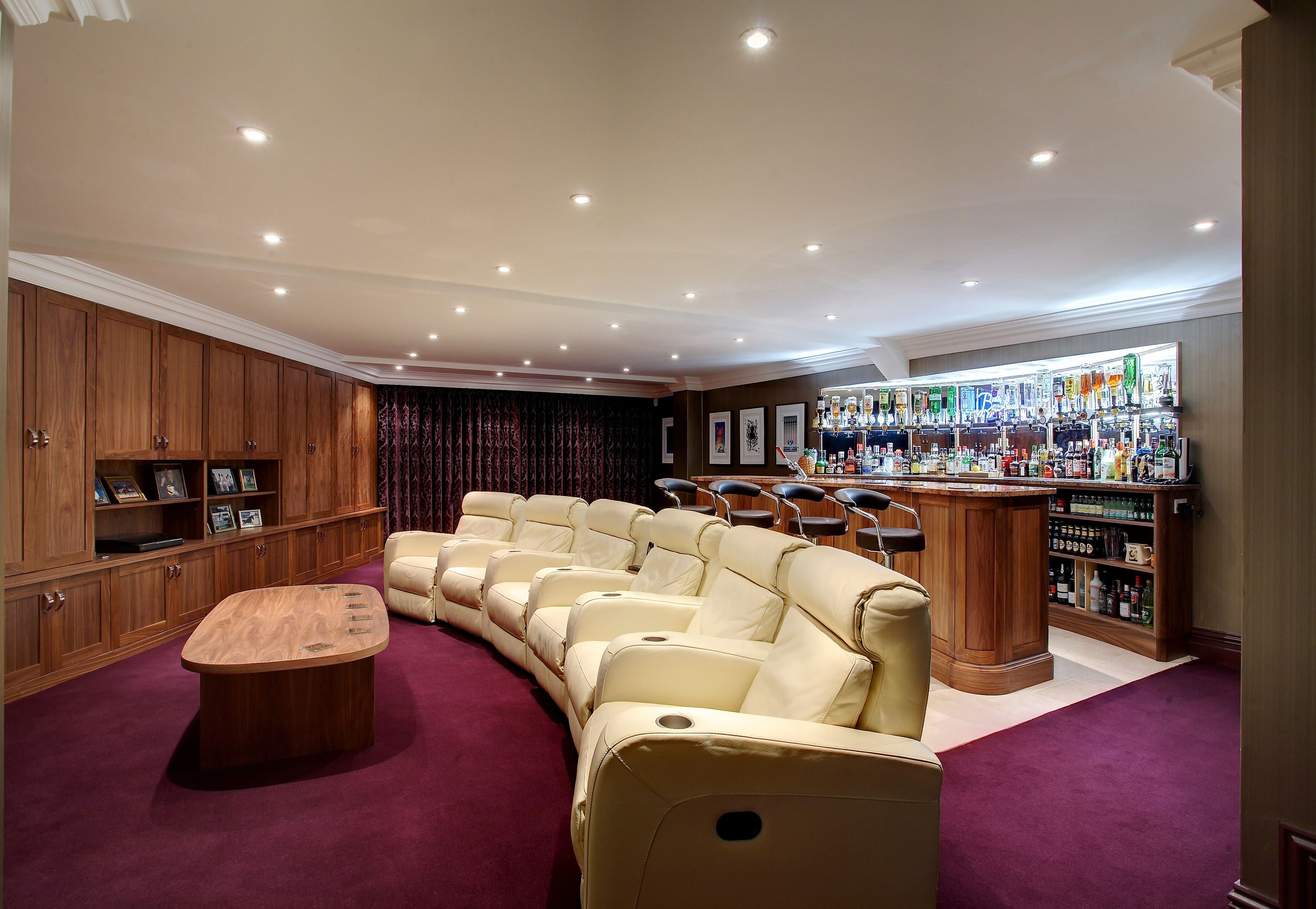 A bespoke, personal cinema room with bold colours. A burgundy carpet leads to a series of cream-coloured leather chairs in a radial shape. The room features floor-to-ceiling wooden cabinets, with a bar across the back wall.