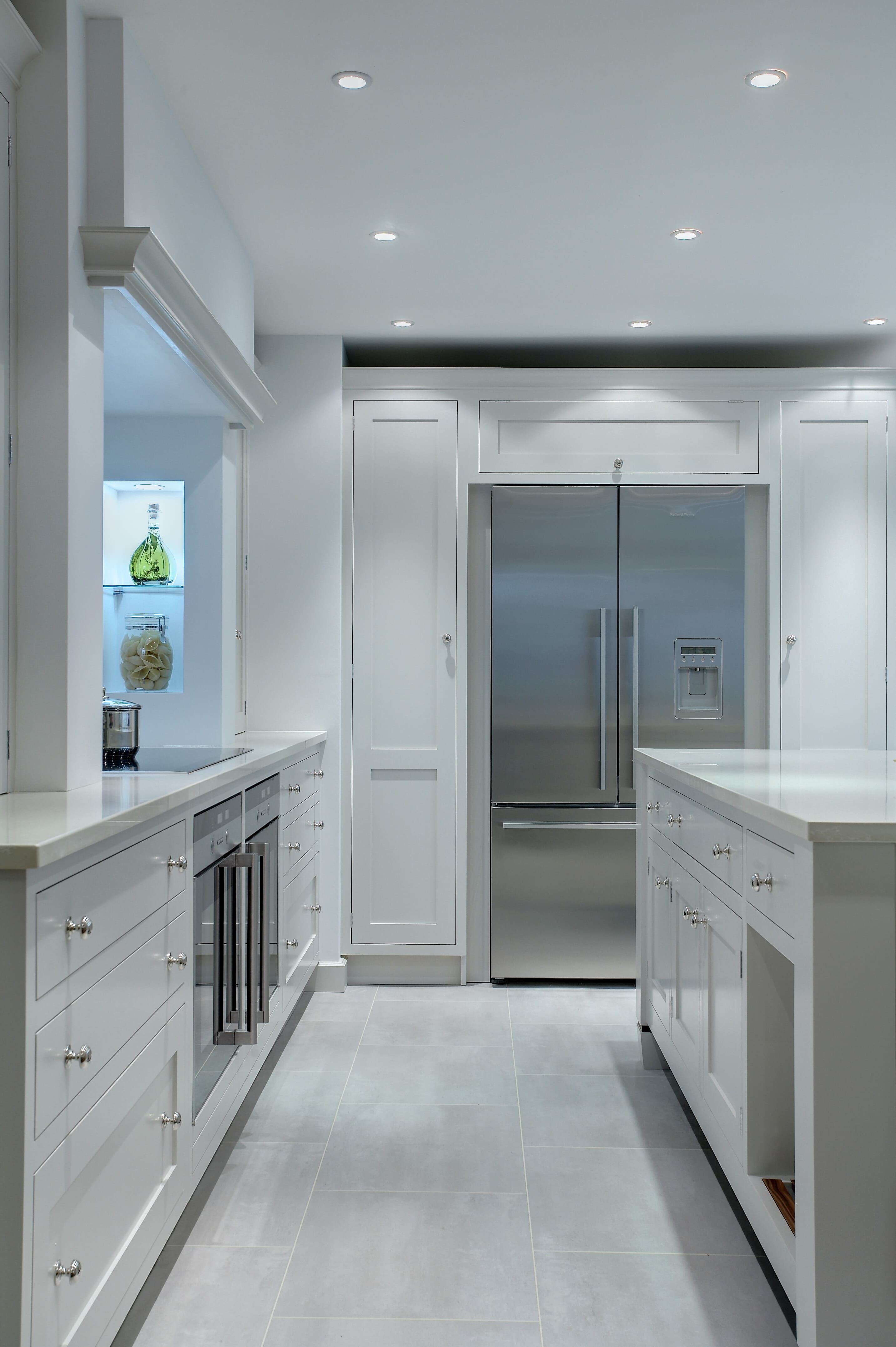 A large, light-grey bespoke kitchen and island is bathed in a cool light from a series of built-in overhead spot lights. A large fridge-freezer can be seen set in to a cabinet on the back wall.