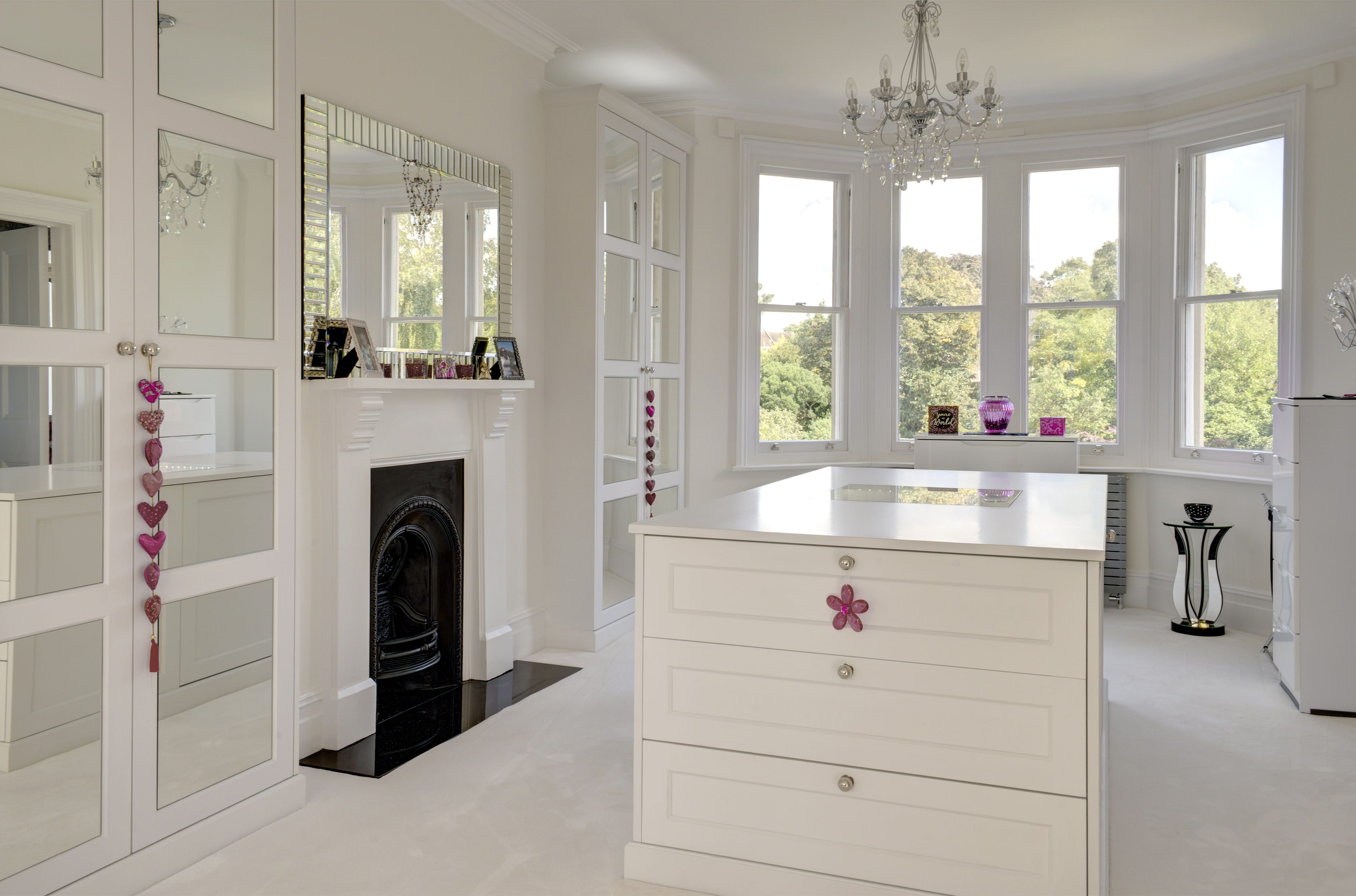A white, bespoke dressing room, with large drawer set/worktop in the centre. Pink hanging decorations provide a contrasting colour.