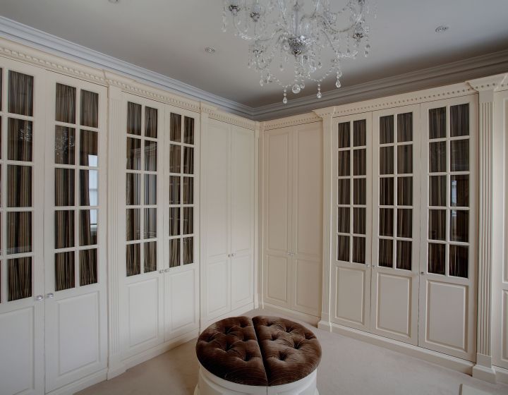 A luxurious bedroom with a neutral colour palette, featuring a large, circular tufted ottoman in the centre. The room has cream fitted wardrobes with paneled doors and glass inserts, offering plenty of storage space. An elaborate crystal chandelier hangs from the ceiling, adding a touch of elegance.